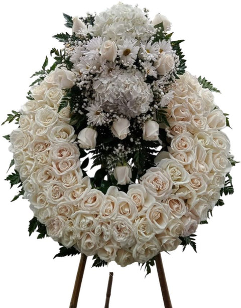 Corona fúnebre circular con abundantes rosas blancas y marfil, flores complementarias blancas y follaje verde, presentada sobre soporte elevado.