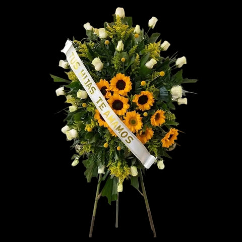 Pedestal funerario de girasoles y rosas blancas sobre soporte elevado, con follaje verde y cinta conmemorativa